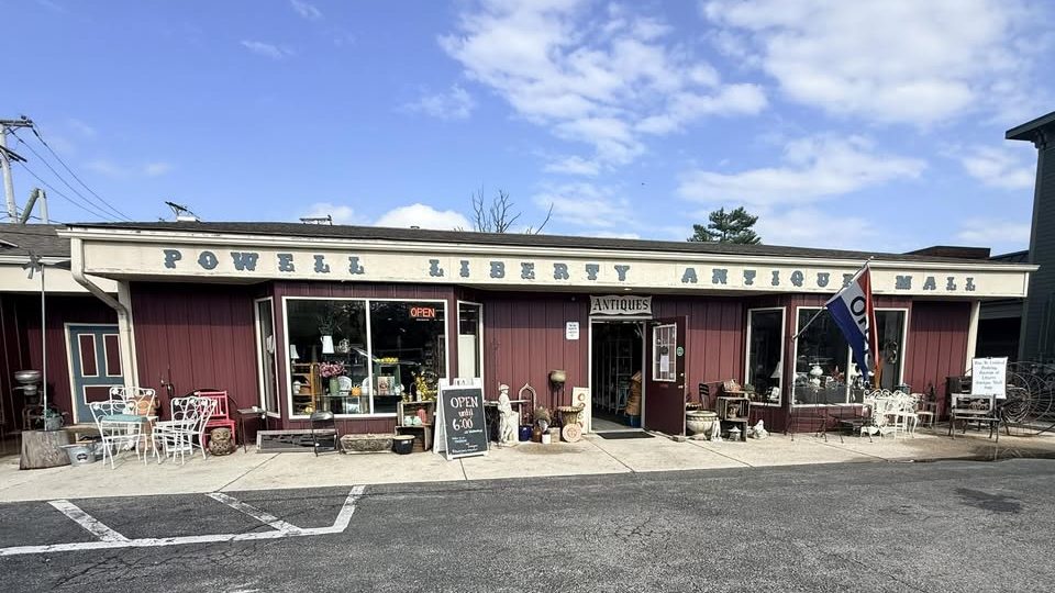 The red storefront of Powell Liberty Antique Mall on a sunny day, with vintage furniture and garden pieces displayed out front